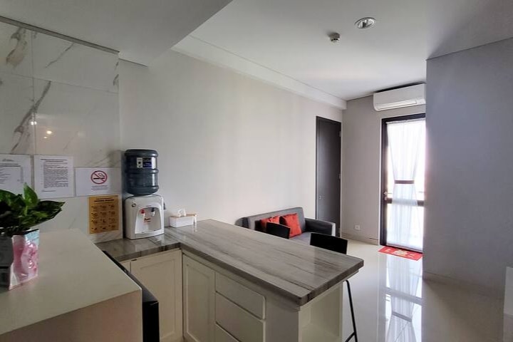The kitchen area features a modern countertop with a water dispenser and potted plant. A small seating area with a black couch and red cushions is visible, along with a door leading to a balcony. Natural light enters through the glass door, brightening the space.