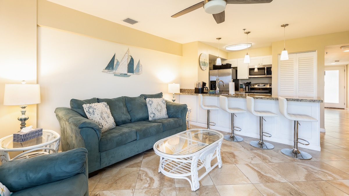 The living area showcases a comfortable teal sofa paired with white wicker seating. A glass coffee table sits on a tiled floor, with a nautical-themed wall decor above. The kitchen features modern appliances and bar seating, illuminated by pendant lighting.