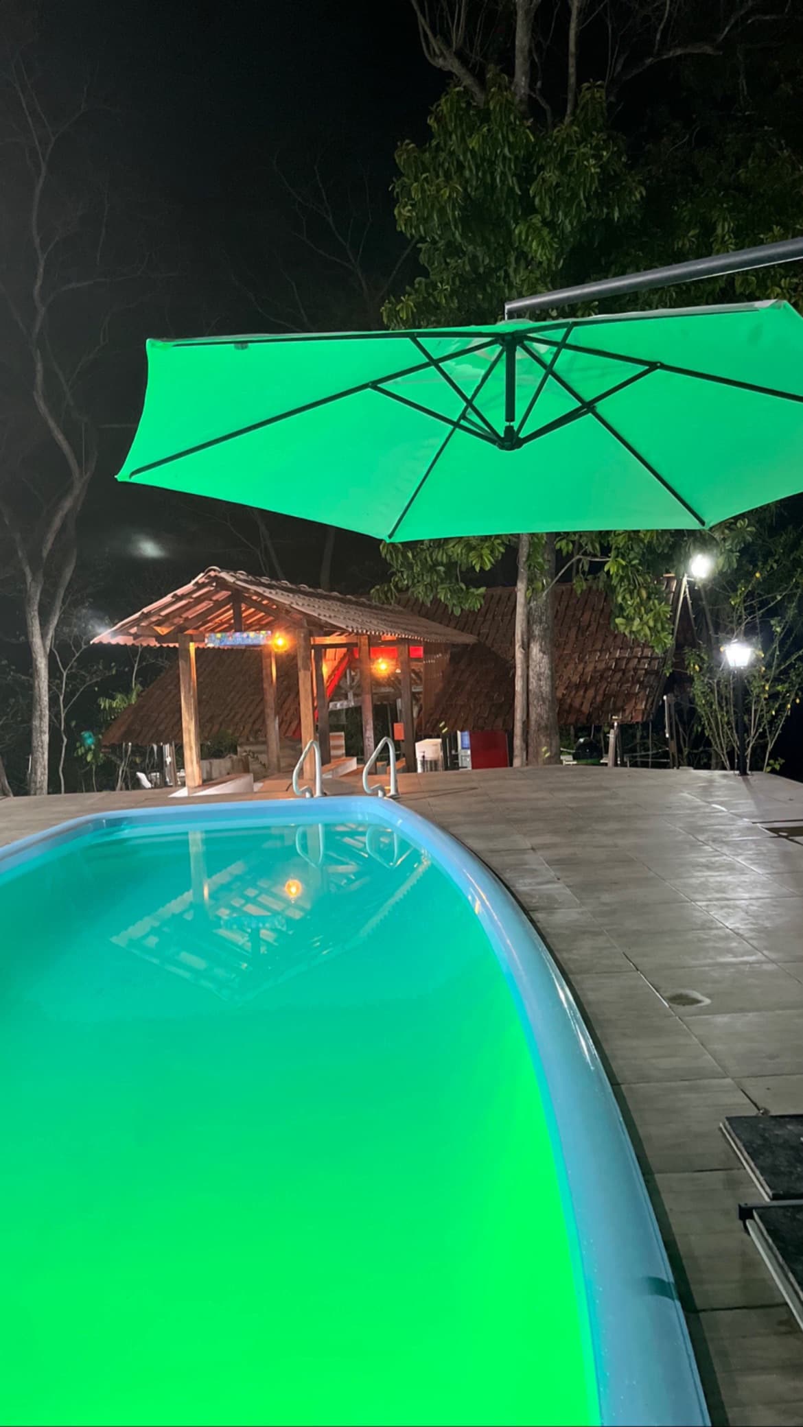 A well-lit swimming pool glows in turquoise tones under a night sky. A green umbrella provides shade beside the pool area, with a wooden structure featuring ambient lighting visible in the background, creating a relaxed outdoor setting.