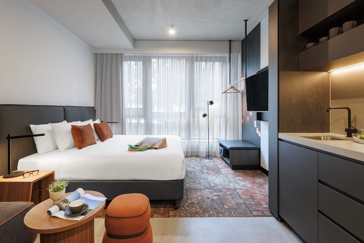 A spacious suite featuring a king-sized bed dressed in crisp linens, complemented by earthy-toned pillows. An inviting lounge area includes a small table and a bright orange ottoman. Large windows allow natural light to fill the room, enhancing the contemporary decor.