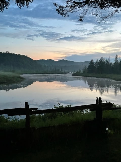 A serene view of the tranquil water reflects the soft colors of the evening sky. Lush greenery frames the scene, with gentle hills in the distance. The stillness of the water and the surrounding nature offer a peaceful ambiance.