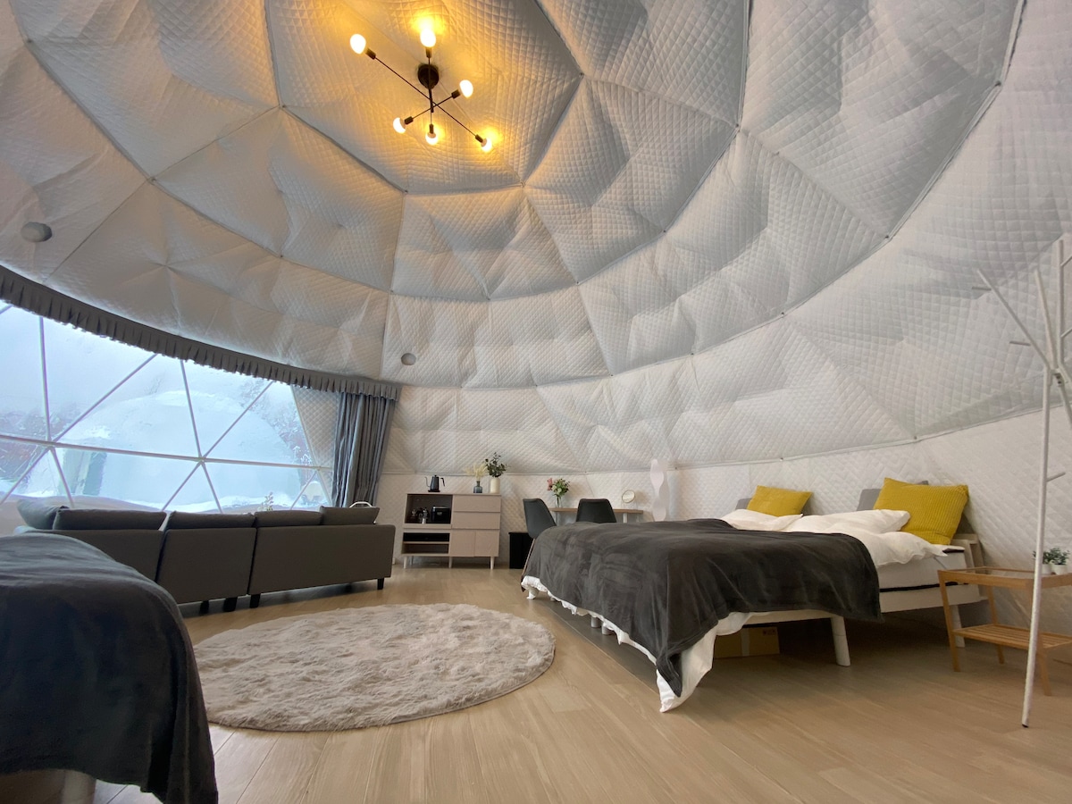 A spacious dome-shaped room features a high, textured ceiling with a modern light fixture. Four single beds are arranged with cozy blankets, alongside a comfortable sofa. Large windows offer a view of the surrounding nature, while a soft rug adds warmth to the wooden floor.