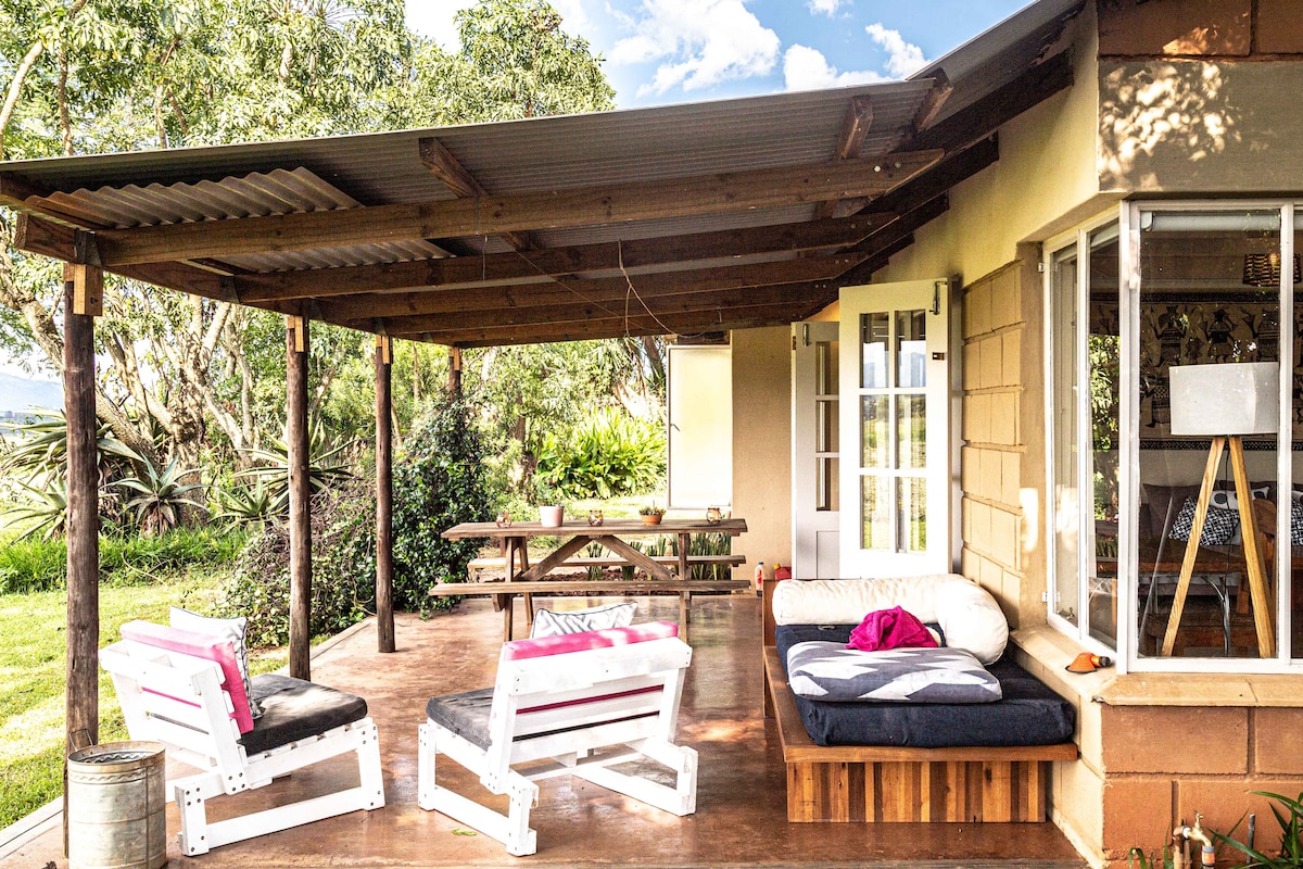 An outdoor patio area features a shaded roof supported by wooden beams. Two white lounge chairs are positioned beside a low wooden platform, which includes cushioned seating. A picnic table with benches sits in the background, surrounded by lush greenery.