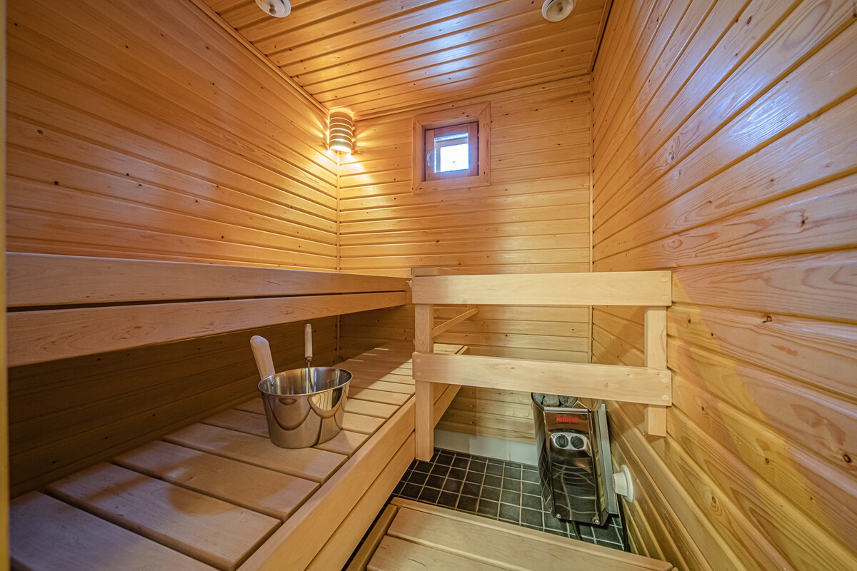 A wooden sauna is showcased with light-colored walls and a ceiling made of wooden planks. Benches run along both sides, with a metal bucket positioned on the lower bench. A small window allows for natural light, while a heat source stands on the floor.
