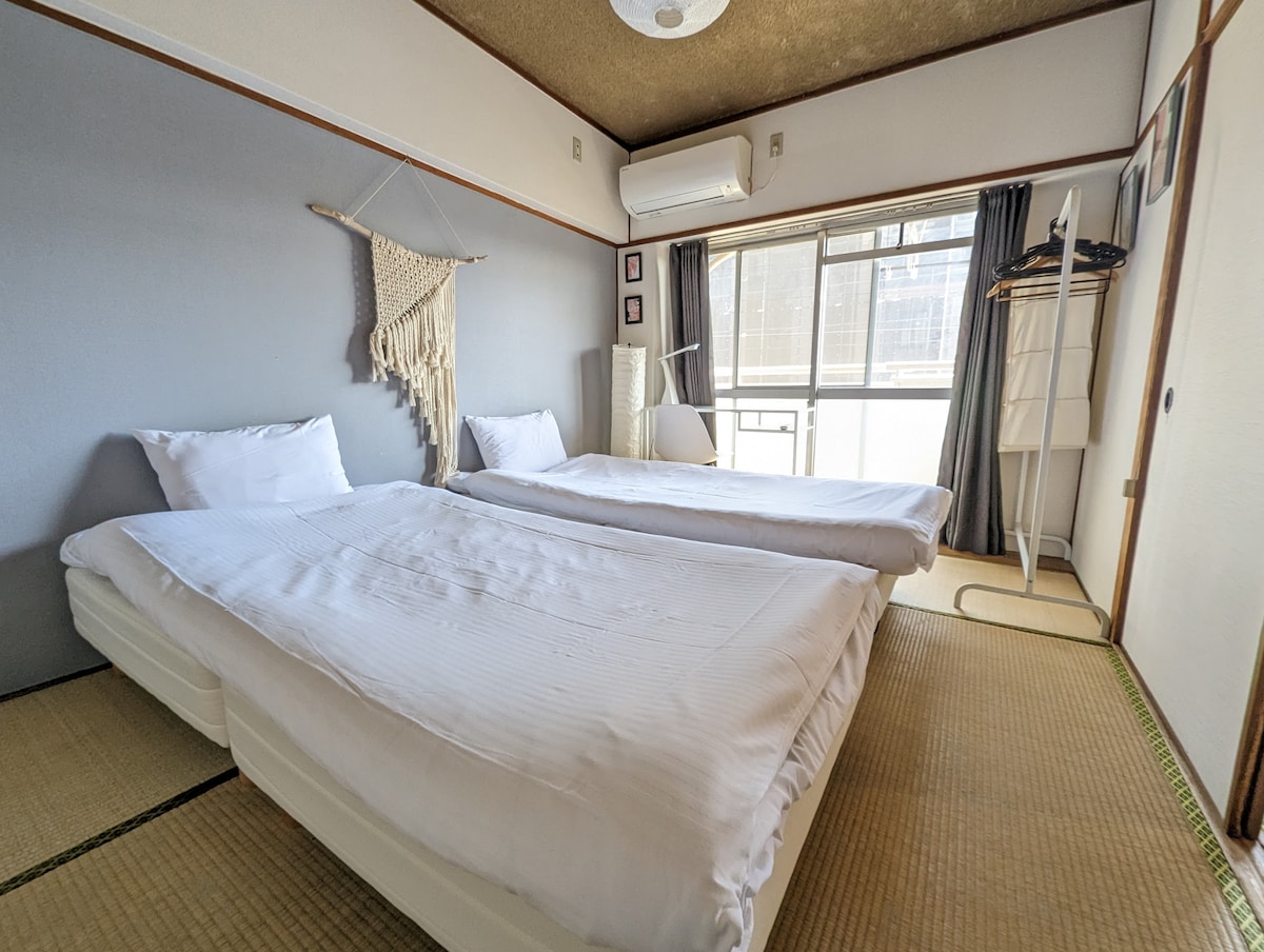 A serene bedroom is equipped with two single beds positioned side by side, featuring crisp white bedding. Natural light filters through a large window adorned with dark curtains. A macramé wall hanging adds a touch of texture, while a wardrobe stands discreetly in the corner.