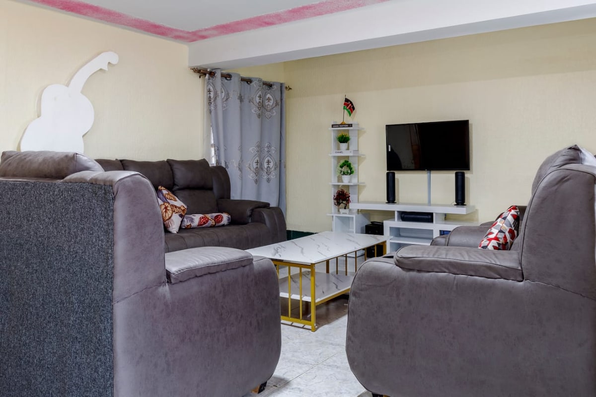 A comfortable living area features soft, plush sofas set around a low coffee table. A mounted flat-screen TV is situated on a white shelving unit, providing an inviting space for relaxation. Light-filtering curtains adorn the window, adding a subtle texture to the room.