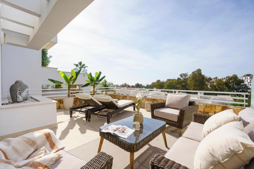 A serene terrace is depicted with comfortable lounge chairs and a small table, surrounded by lush greenery. The space features an outdoor seating arrangement with plush cushions, while a cozy blanket rests on one chair, inviting relaxation and enjoyment of the sunshine.