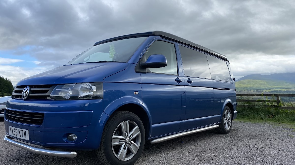 A blue Volkswagen campervan is parked on a gravel road. The vehicle features a sleek design with chrome accents, large windows, and a roof that enhances the spacious feel. Lush green hills and a cloudy sky can be seen in the background.