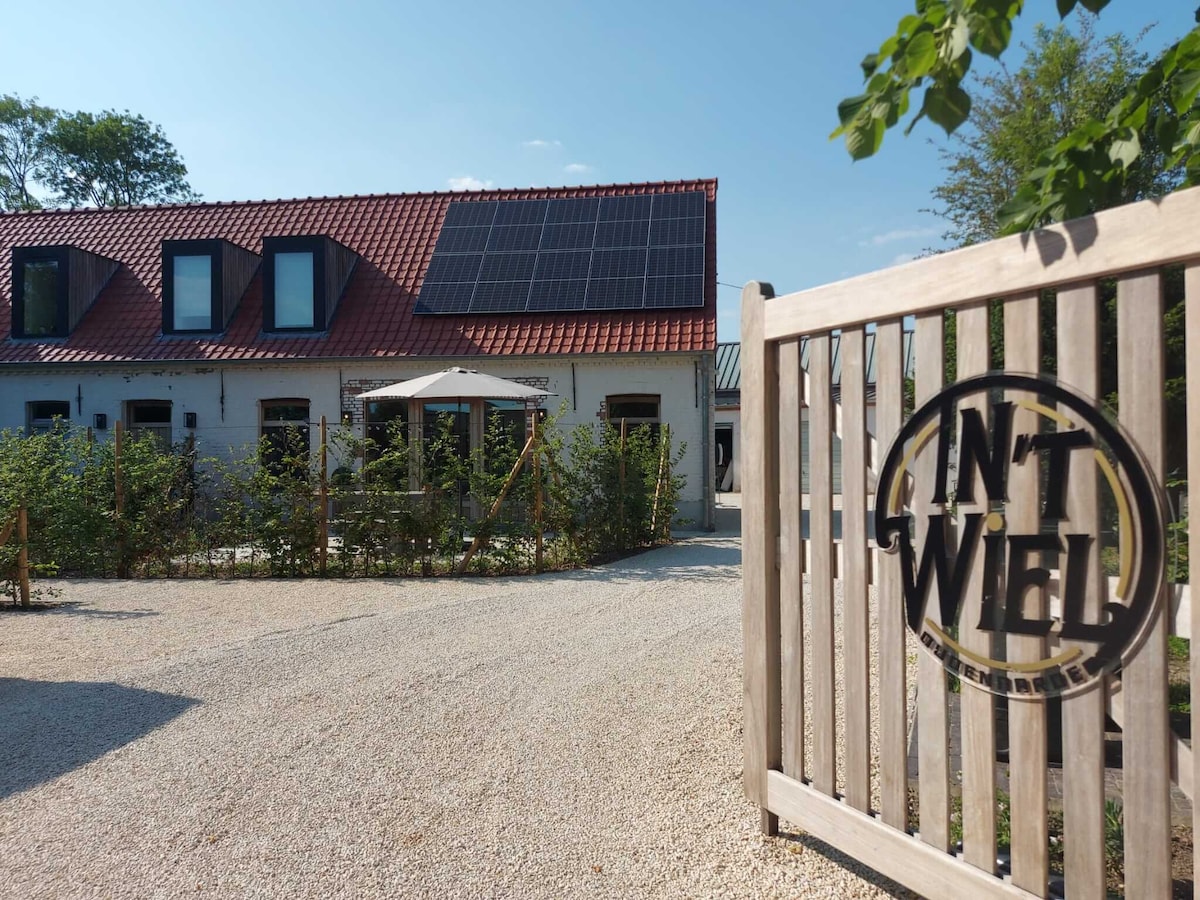 The exterior of the vacation home is shown, featuring a gravel driveway leading to a welcoming entrance. Solar panels are installed on the red-tiled roof. A wooden gate with the name 'In ’t Wiel' is prominently displayed, surrounded by greenery.