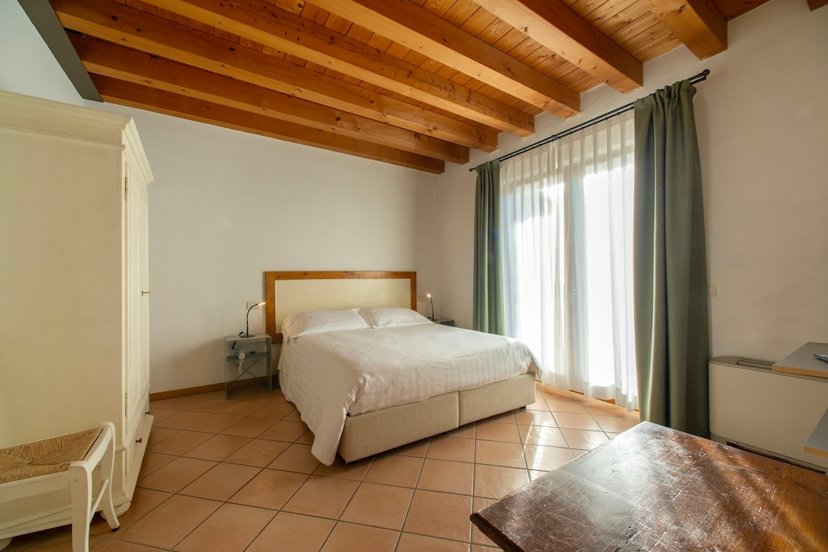 A spacious sleeping area is presented with a large bed featuring a light-coloured headboard against a backdrop of exposed wooden beams. Natural light enters through large glass doors draped with sheer curtains, illuminating the tiled floor. A minimalist wooden table and wardrobe are included in the room.