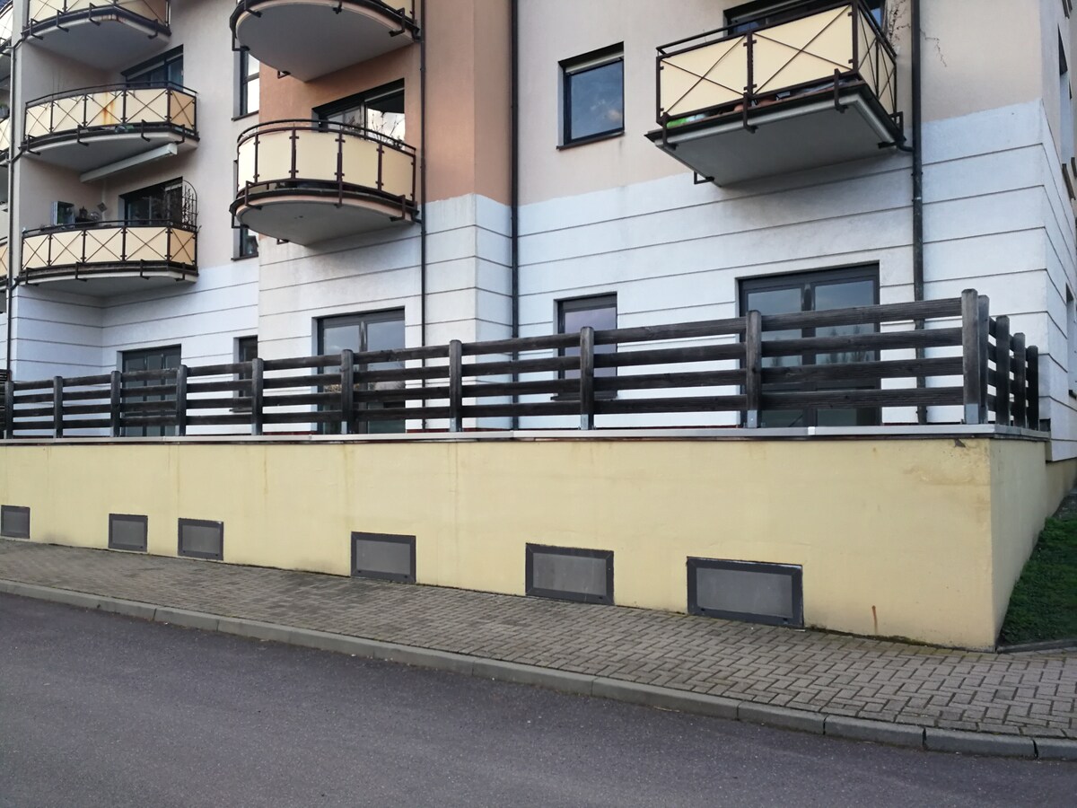 A modern building exterior features a light-colored wall complemented by dark wooden fencing. Three balconies are visible above, each with metal railings. Windows are framed with dark accents, and the area is accessible from the street, showcasing ample space for outdoor relaxation.