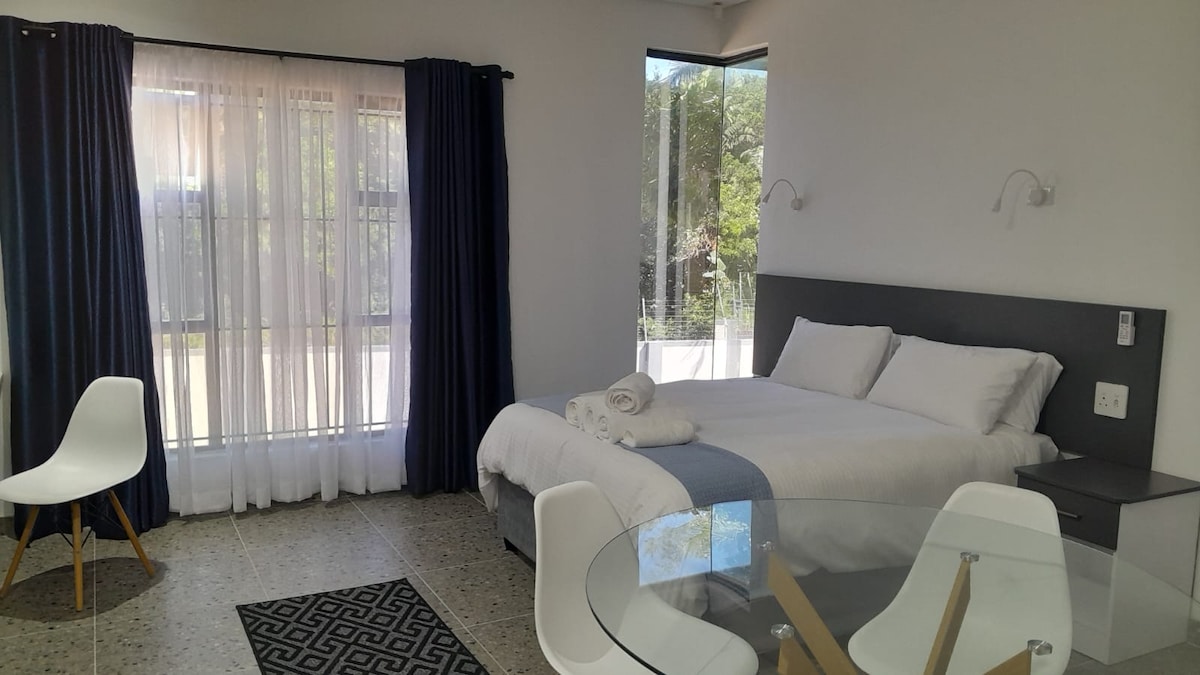 A spacious bachelor unit features a comfortable bed with fresh linens, complemented by a neutral color scheme. A dining table with clear chairs is present, alongside large windows draped in dark curtains that allow natural light to fill the room.