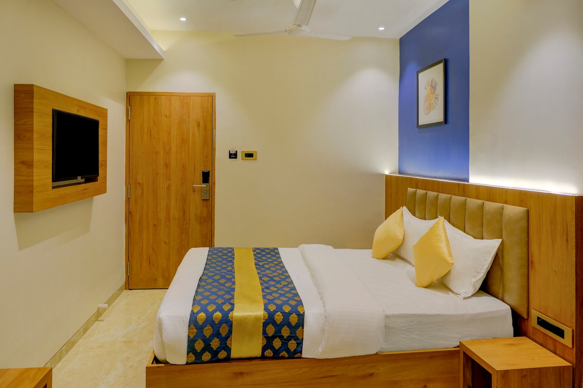 A well-lit room is showcased with a spacious bed featuring a blue and gold decorative runner and two yellow accent pillows. A wooden TV unit is mounted on the wall across from the bed, and a wooden door leads to the hallway.