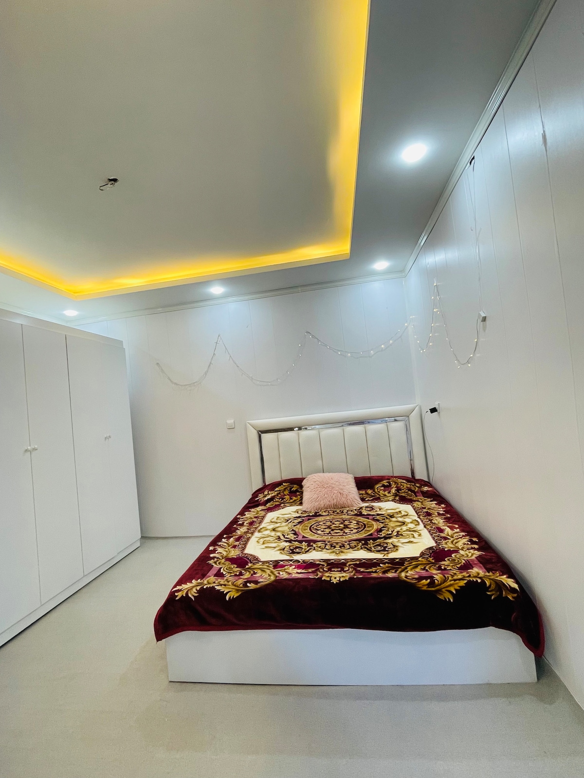 A bright sleeping area features a spacious bed adorned with a richly patterned blanket. Soft lighting is provided by recessed ceiling lights, and white cabinetry lines one wall. Decorative string lights create an inviting ambiance within the room.