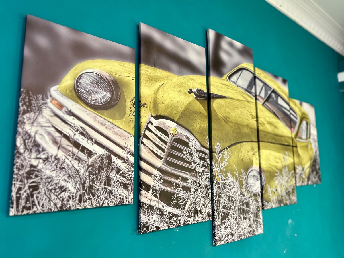 A collection of five canvas panels displays a vintage yellow car partially obscured by tall grass, set against a bright turquoise wall. The artwork captures detailed features of the car, including its iconic grille and headlights.