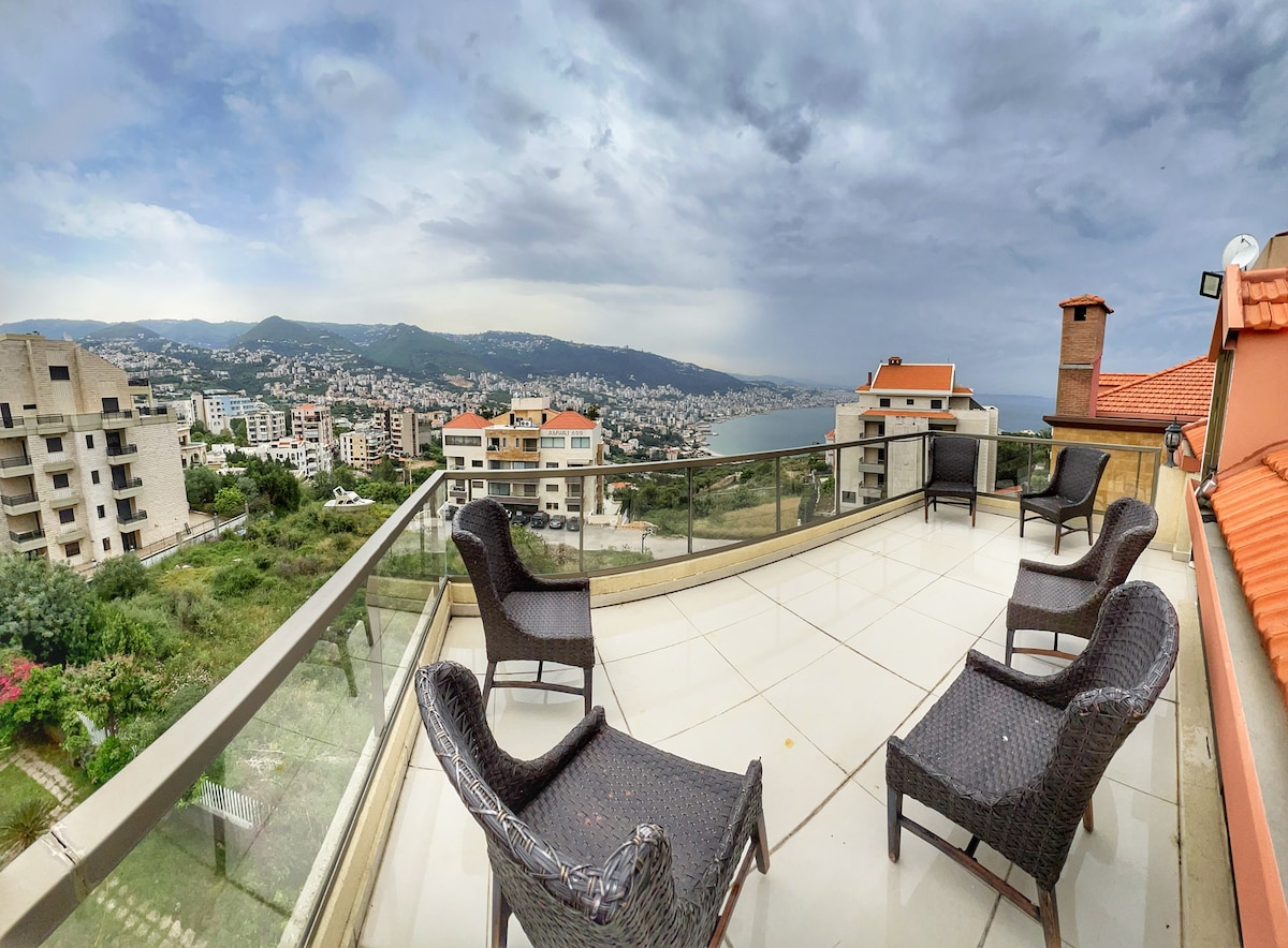A spacious balcony with a glass railing offers an unobstructed view of the coastline and mountains. Four wicker chairs are arranged around the area, providing a comfortable setting for relaxation. The surrounding landscape features a mix of residential buildings and greenery, under a cloudy sky.