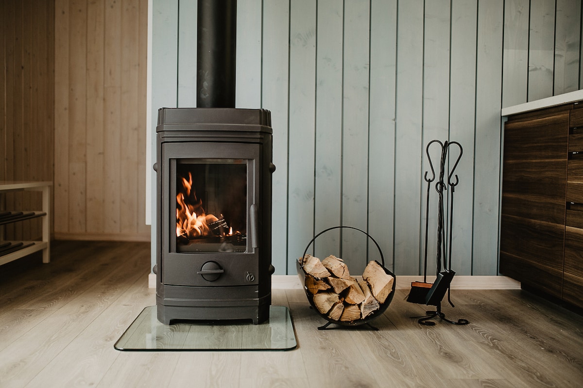 A modern wood-burning stove is positioned against a light blue wooden wall, emitting a warm flame. Beside it, a cylinder of neatly stacked firewood rests in a metal holder, with a black set of fireplace tools nearby.