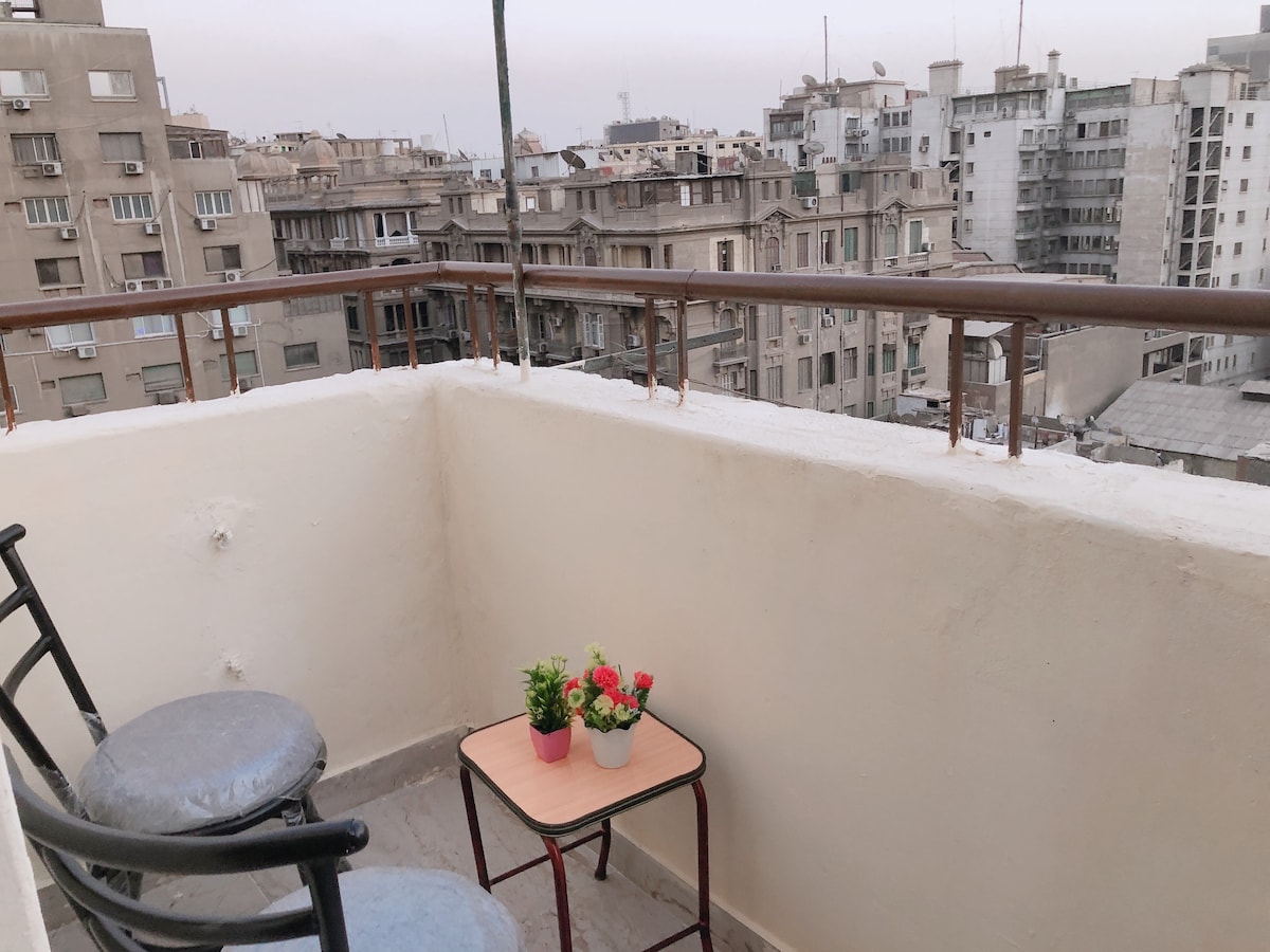 A small balcony features two black chairs and a compact table. A potted plant with colorful flowers sits on the table. The view reveals a mix of historical and modern buildings in the distance, showcasing the urban landscape.