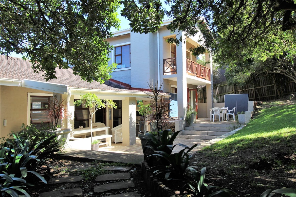 The exterior of the property reveals a two-story home surrounded by lush greenery. A staircase leads to a balcony, while a patio with seating areas is visible on the ground floor. The entrance is framed by trees, providing a natural, shaded environment.