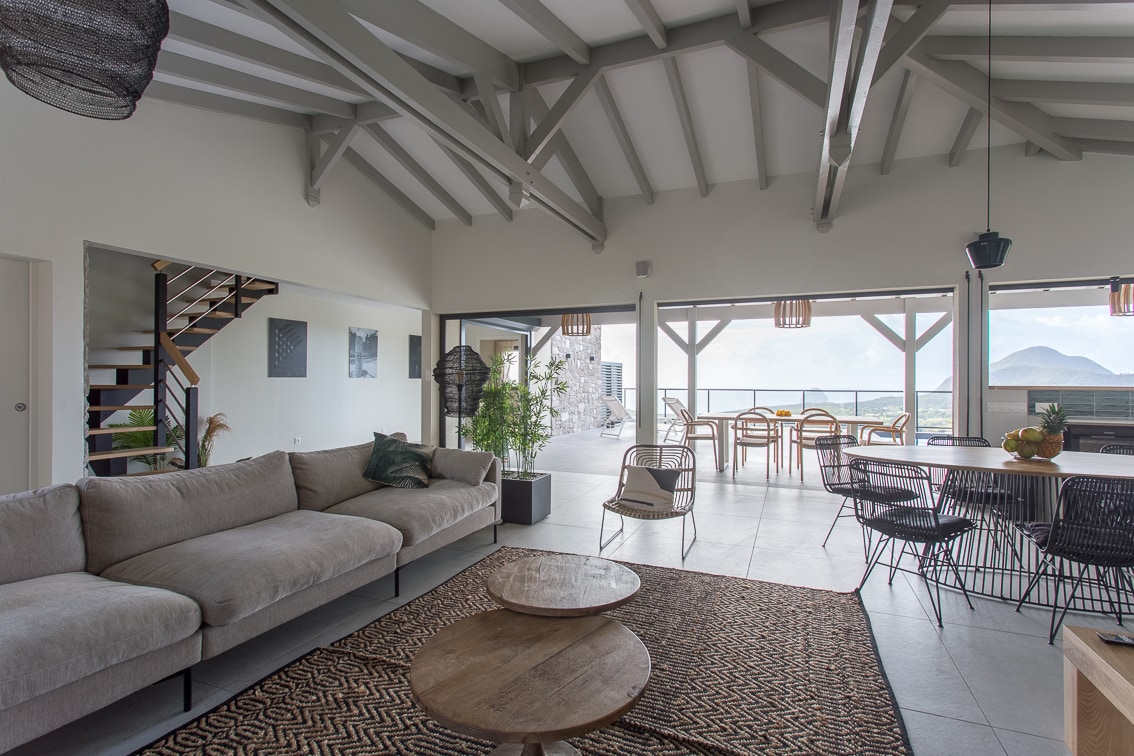 An open-concept living area is highlighted by a spacious sectional sofa on a textured rug, positioned across from a wooden coffee table. Large glass doors reveal a stunning view of the surrounding landscape, while a dining area with wooden chairs and a table is visible nearby.