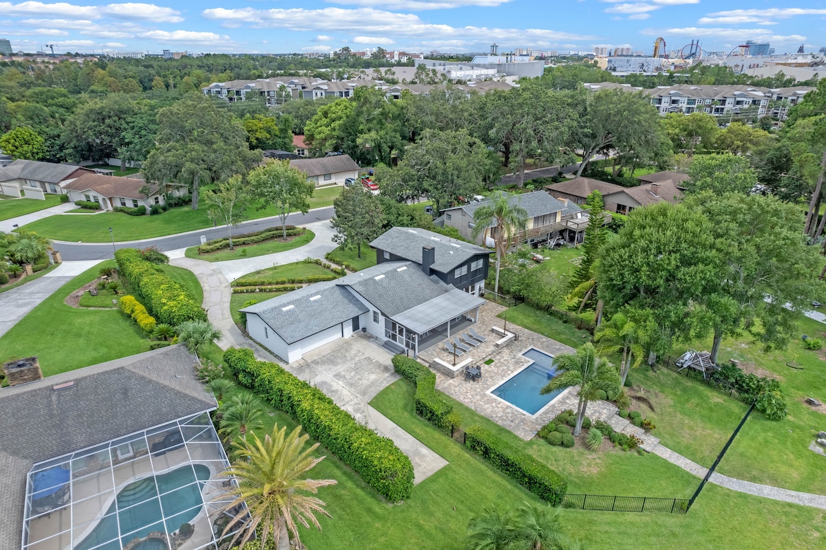 An aerial view of the property highlights the spacious backyard featuring a serene pool surrounded by paved areas and lush greenery. The multi-level home stands prominently alongside well-maintained landscaping, while neighboring homes and community amenities are visible in the distance.