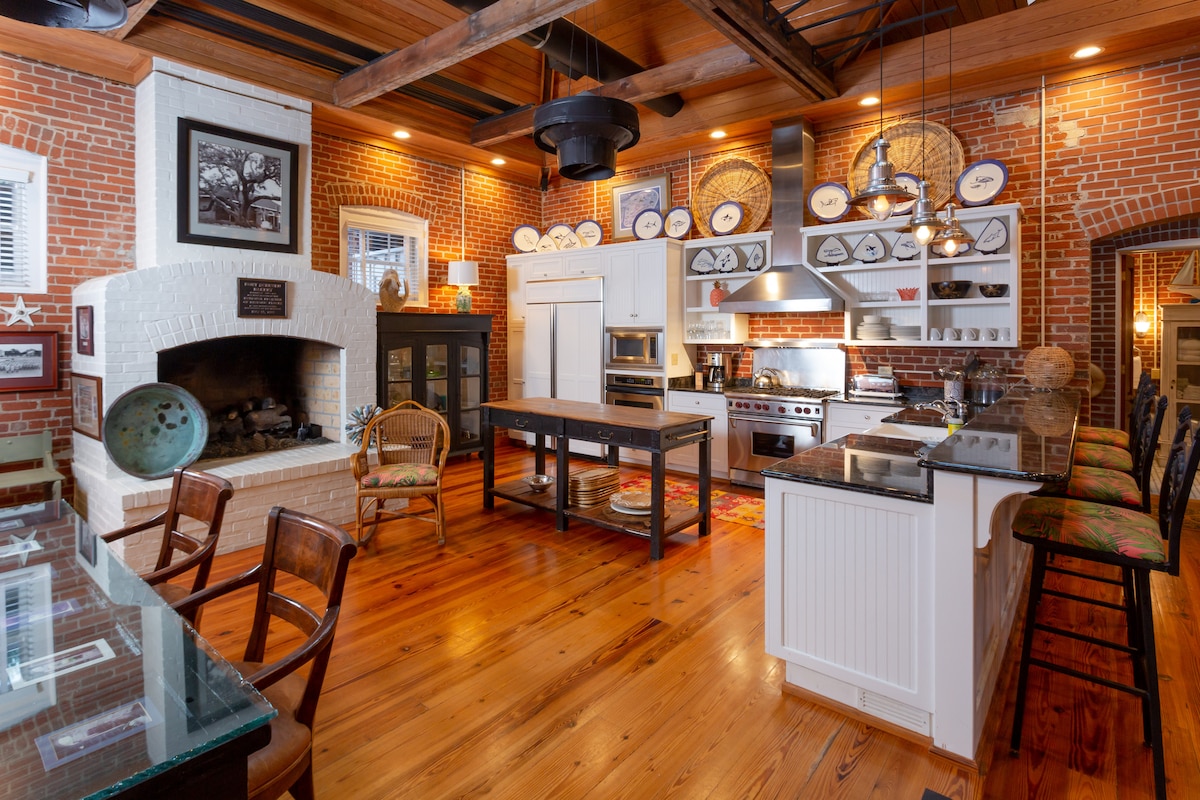 The kitchen features exposed brick walls and wooden beams, enhancing its rustic charm. A large kitchen island stands at the center, complemented by bar seating. High-end appliances and decorative dishware can be seen, while a cozy fireplace adds warmth to the inviting space.