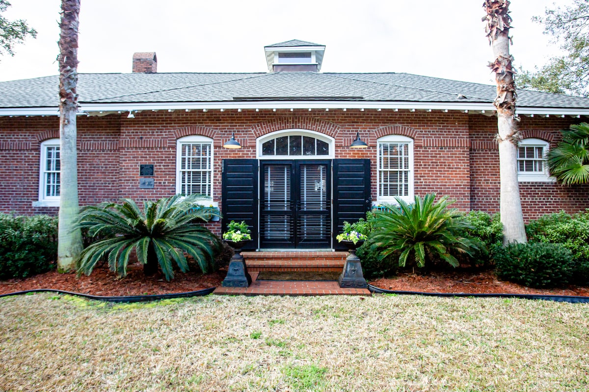 The exterior of The Bakery showcases a historic brick façade, framed by palm trees and lush greenery. Black shutters are closed over the windows, and a welcoming front entry with double doors is visible. A neatly maintained lawn complements the inviting atmosphere.