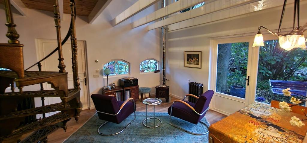 A spacious living area features two purple chairs positioned around a small table, accompanied by a colorful bedspread. Large windows illuminate the space, offering views of the lush garden outside. An ornate spiral staircase adds architectural interest, while a vintage radio sits on a nearby shelf.