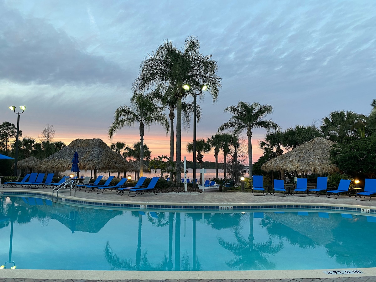 A serene pool area is surrounded by palm trees and tiki huts, reflecting the colorful twilight sky. Lounge chairs in vibrant blue are positioned around the pool, creating a relaxing setting for guests to unwind.