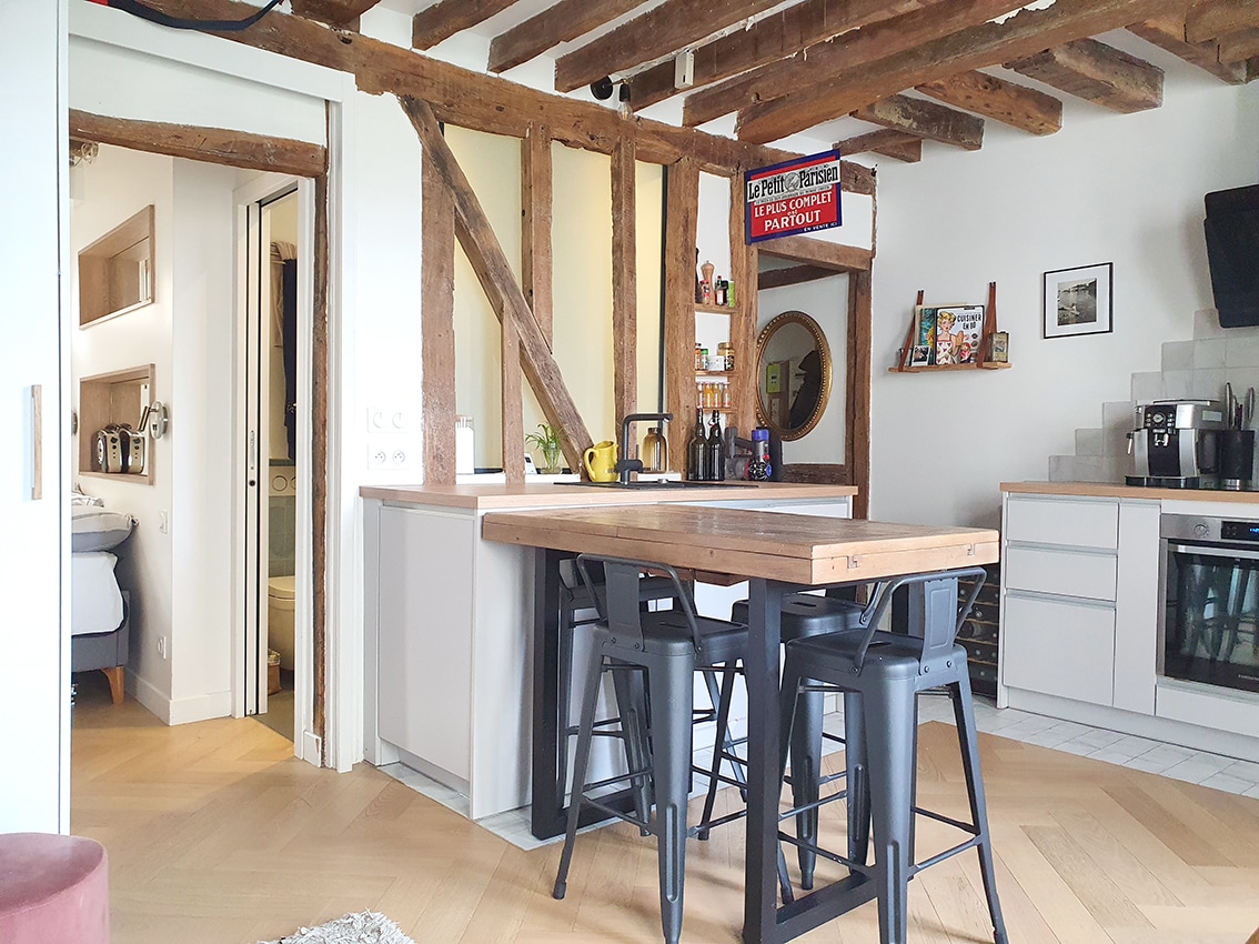 A modern kitchen features an island with wooden countertops surrounded by black stools. Exposed wooden beams enhance the ceiling. A mirror and decorative items are positioned on the wall, while kitchen appliances are neatly arranged on the counters in a functional layout.