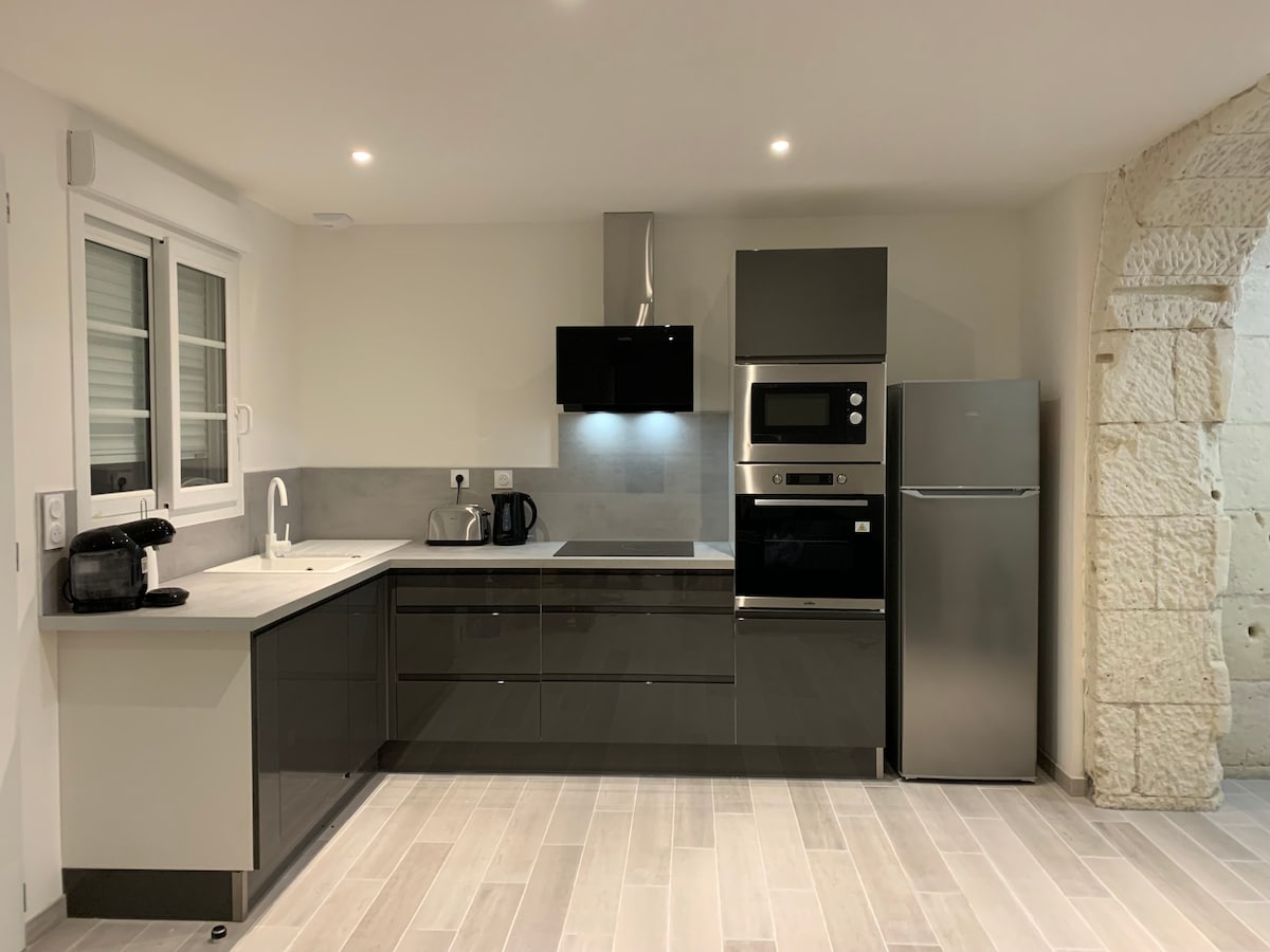The kitchen features modern cabinetry with a sleek finish, complemented by a dark countertop. Appliances include a refrigerator, oven, microwave, and kettle. A sink is positioned under a window, allowing for natural light. The room showcases a clean and functional design with neutral tones.