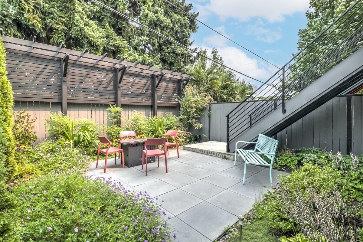 A private patio is featured with a stone surface, surrounded by lush greenery and colorful plants. Outdoor furniture includes a table with four chairs and a light blue chair, all positioned under a wooden pergola, providing a tranquil space for relaxation.