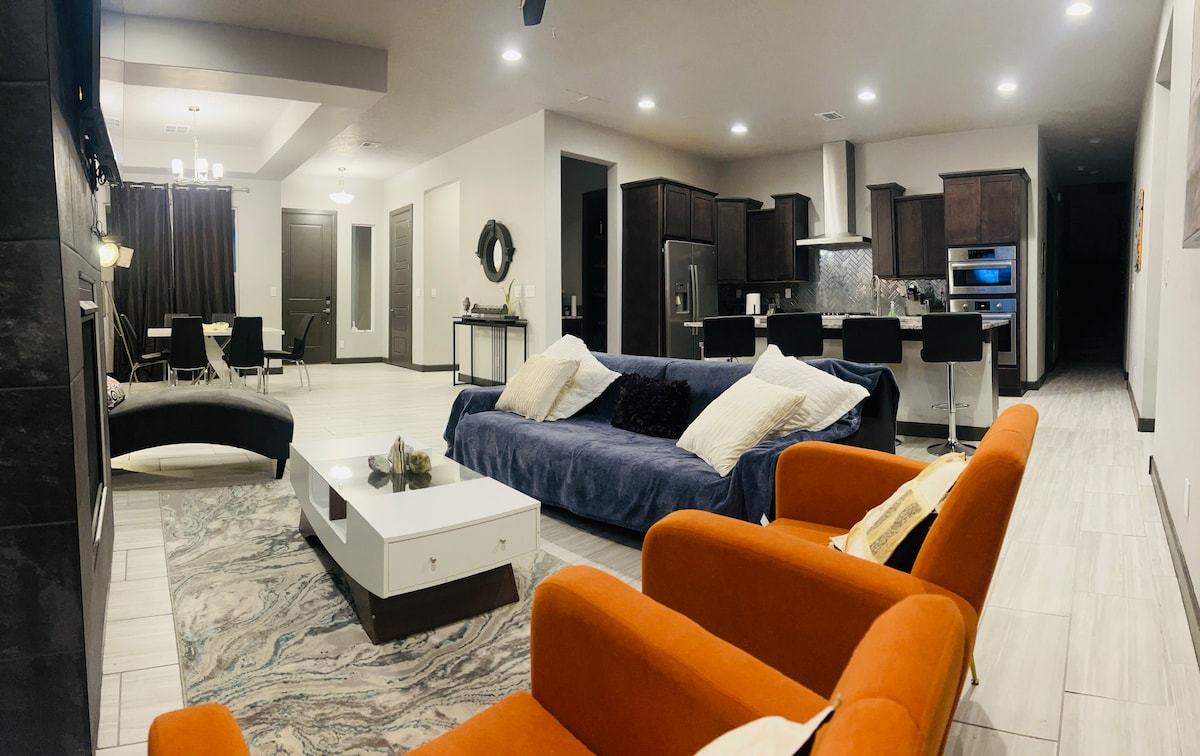 The spacious open-plan living area features plush seating in orange and blue hues. A modern coffee table sits in front of a sleek entertainment unit. Behind, the kitchen showcases dark cabinetry and stainless steel appliances, with a dining area visible in the background.