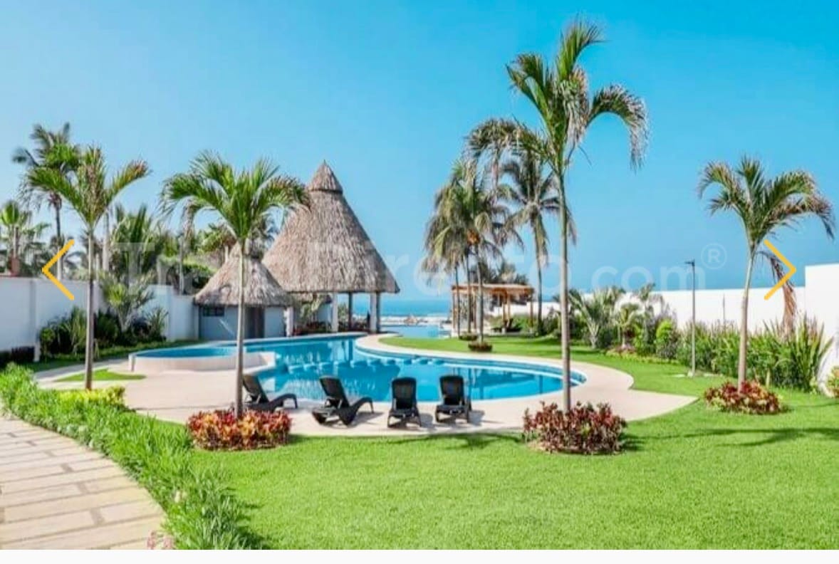 A spacious outdoor area is featured, showcasing a large swimming pool surrounded by manicured gardens. Palm trees provide shade, and thatched-roof structures are positioned nearby. Comfortable lounge chairs are arranged around the pool, emphasizing a relaxed atmosphere.