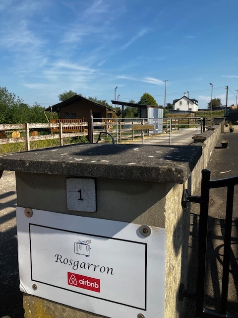 A welcoming entrance is marked by a stone wall featuring a sign labeled 'Rosgarron.' Beyond the wall, the area reveals rustic charm, with a gravel path leading toward lodges and green landscapes, set against a clear blue sky.