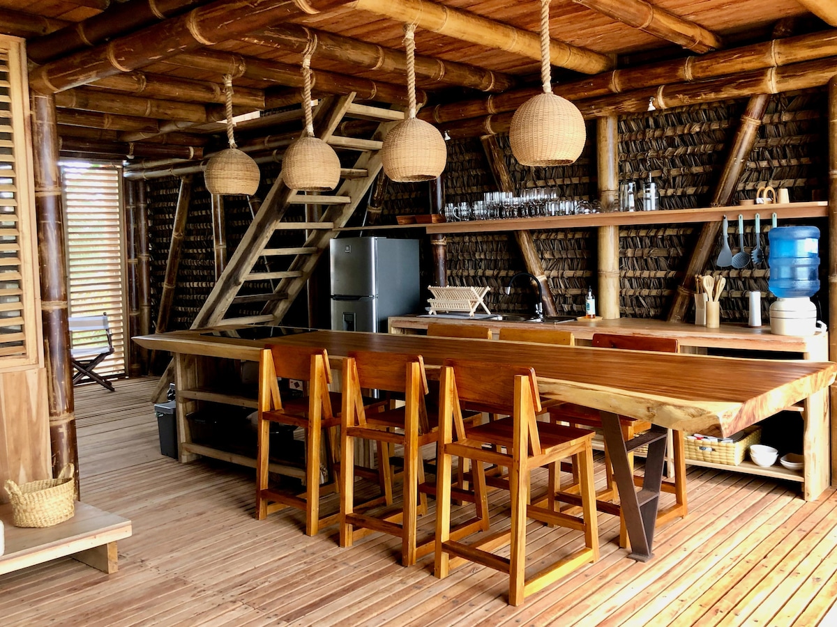 An open kitchen area features a large wooden table paired with sturdy seats. Natural materials dominate the design, with bamboo beams supporting the structure. A staircase leads to an upper level, and various kitchen items are neatly arranged on shelves above a modern refrigerator.