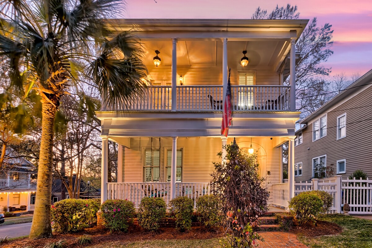 A charming two-story home features a welcoming front porch adorned with white railings and elegant lighting. Lush landscaping surrounds the entrance, with vibrant flowers and palm trees contributing to the inviting exterior. The setting sun casts a warm glow, highlighting the Southern architectural details.