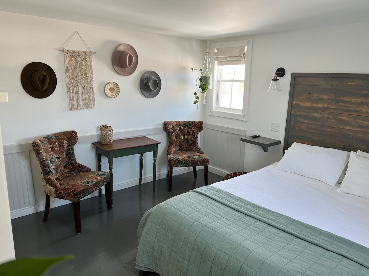 A guest room features a spacious bed with a light-colored quilt. Two upholstered chairs with floral patterns are positioned beside a small table. Wall-mounted accents include natural materials, decorative hats, and a hanging textile, enhancing the room's charm and character.