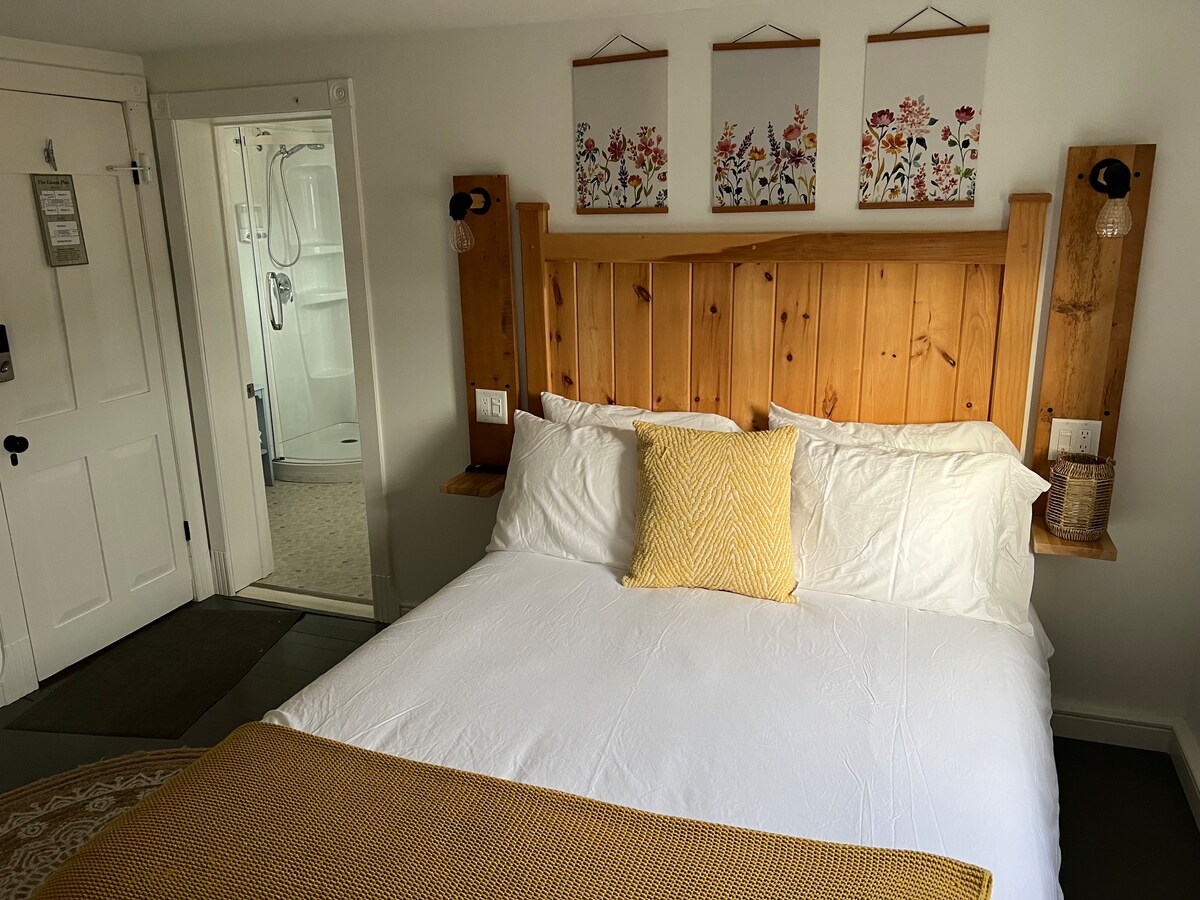 A queen size bed is framed by wooden headboard accents, with a yellow decorative pillow resting on the white linens. Floral art pieces hang above the bed, while a private ensuite is visible in the background, featuring a walk-in shower and neutral tones.