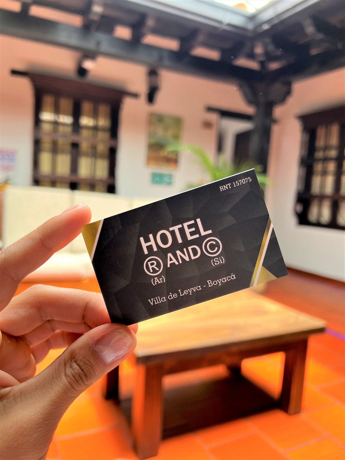 A hand holds a card displaying the hotel's name and location. In the background, a cozy common area is visible, characterized by terracotta tiles and wooden furniture. Large windows allow natural light to fill the space, providing a welcoming ambiance.