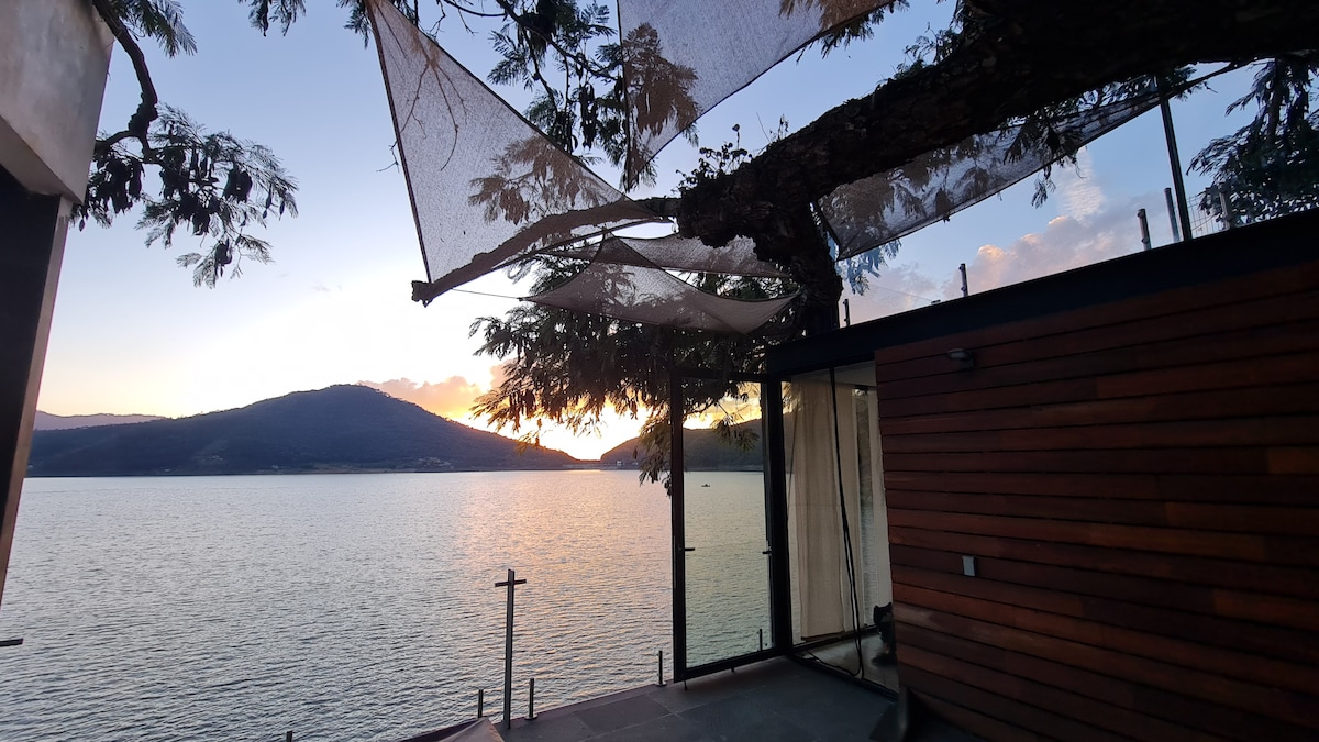 An outdoor terrace features a natural canopy formed by tree branches, while soft fabric panels provide shade. The view reveals a tranquil lake, with mountains in the background illuminated by the warm colors of sunset. The serene atmosphere invites relaxation.