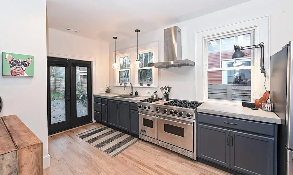 The kitchen features dark cabinetry with stainless steel appliances, including a professional-grade oven. Natural light enters through multiple windows, illuminating the countertop. A dining area is accessible through a set of black doors, while a striped rug adds texture to the wooden floor.