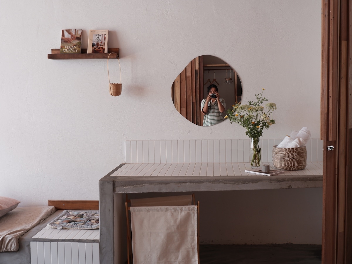 A minimalist workspace features a circular mirror above a concrete table. Fresh flowers are arranged in a woven basket, adding a touch of nature. A light-colored chair is positioned at the table, while a cozy bed and magazines are visible in the background.