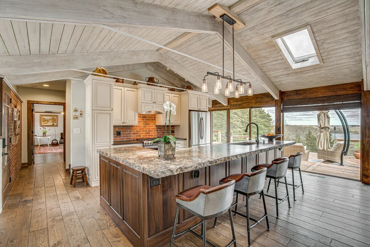 An open kitchen area features a large island with seating and granite countertops. Light wood beams accent the vaulted ceiling, while skylights allow natural light to fill the space. Transom windows offer views of the outside, enhancing the inviting atmosphere.