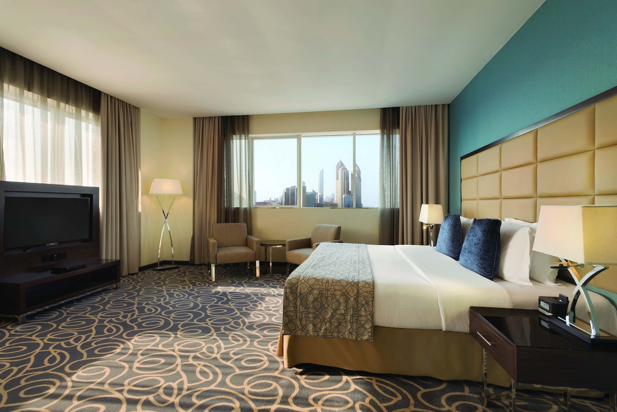 A comfortable hotel room is featured, showcasing a spacious bed with decorative pillows against a textured wall. Natural light enters through large windows, illuminating seating areas with two armchairs. A flat-screen television is positioned on a dark wooden console, enhancing the modern aesthetic.