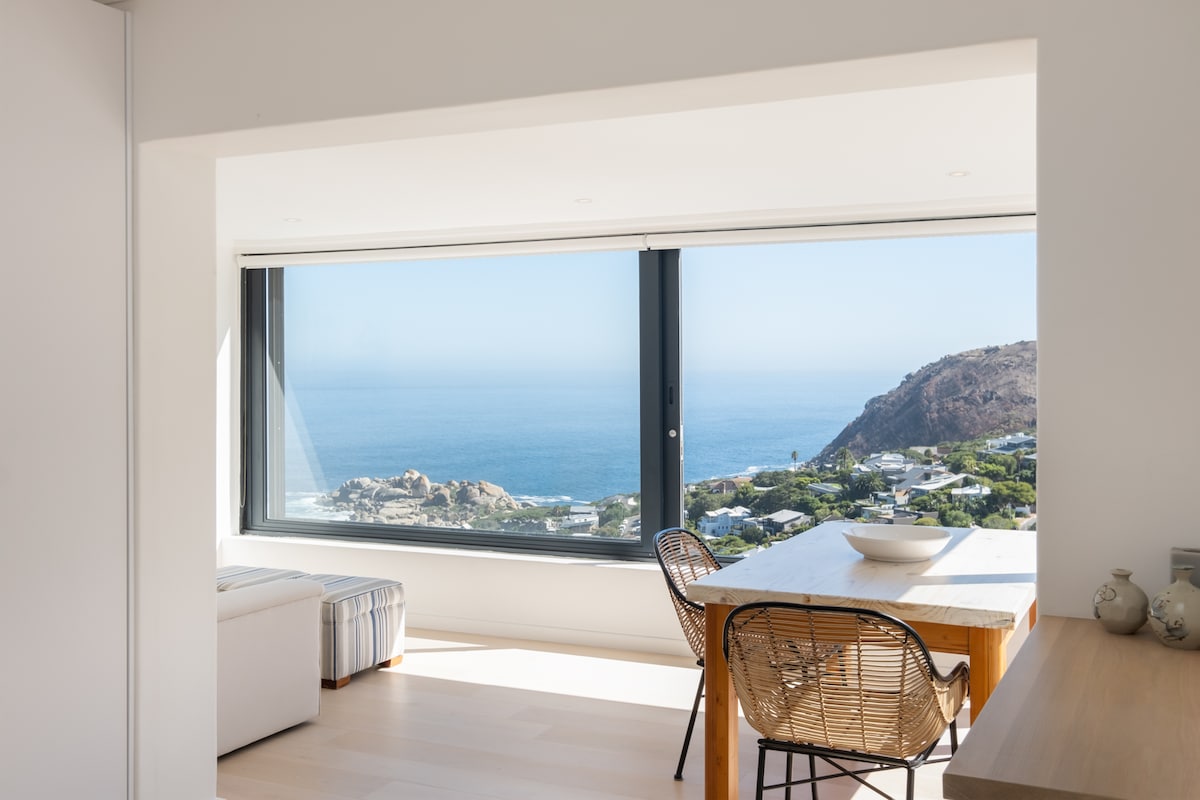A spacious dining area features a wooden table paired with stylish chairs, bathed in natural light. A large window offers unobstructed views of the ocean and coastal landscape, creating a serene atmosphere. Soft textures and a neutral palette enhance the modern design.
