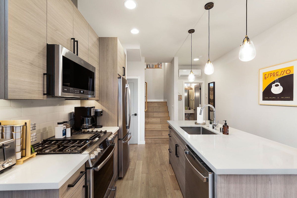 The kitchen features modern appliances including a gas stove, microwave, and refrigerator, complemented by wooden cabinetry. A large island provides additional counter space, while pendant lights illuminate the area. The clean layout is enhanced by natural wood flooring and a view towards the living area.