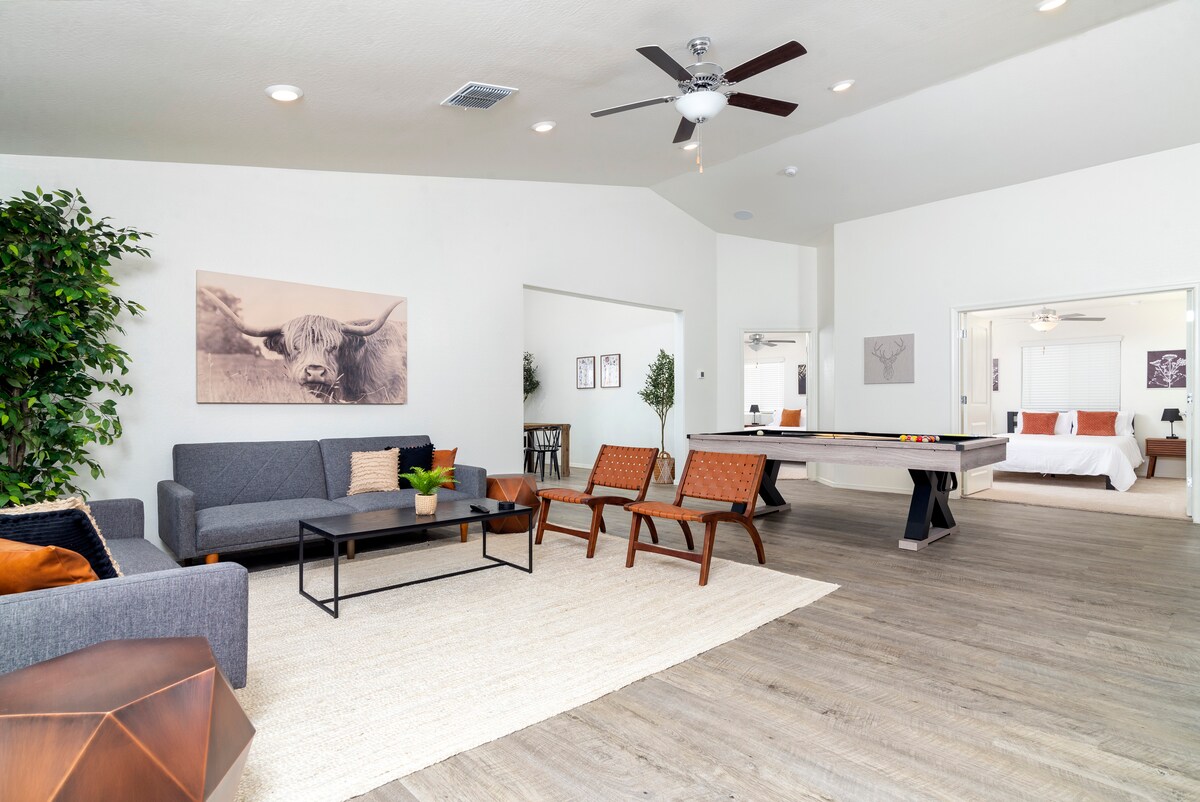 The open-concept living area showcases a comfortable seating arrangement with two sofas and accent chairs, a coffee table, and a pool table. Natural light fills the space, complemented by modern artwork on the walls and a ceiling fan for added comfort.