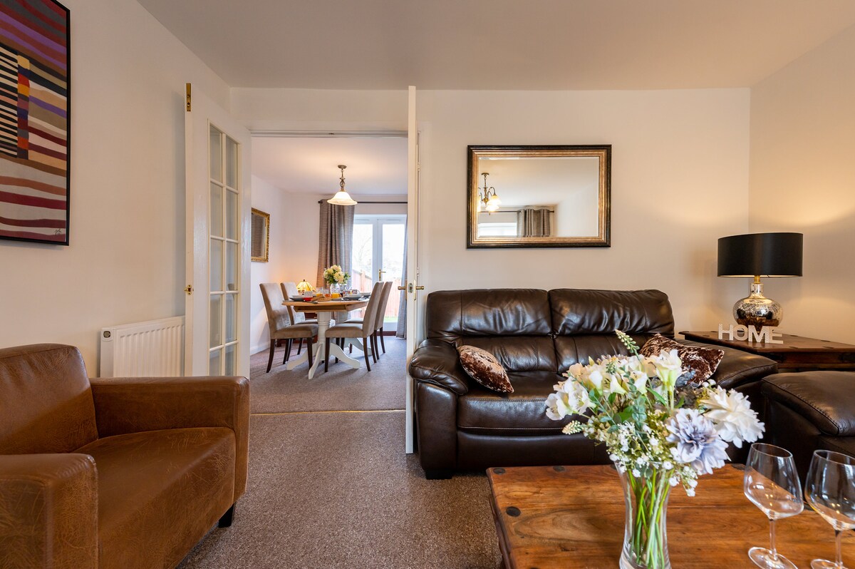 A comfortable lounge area features dark leather sofas and a wooden coffee table, accented by a vase of fresh flowers. A mirror reflects the light from the adjoining dining room, where a table is set for a meal, visible through a set of double doors.