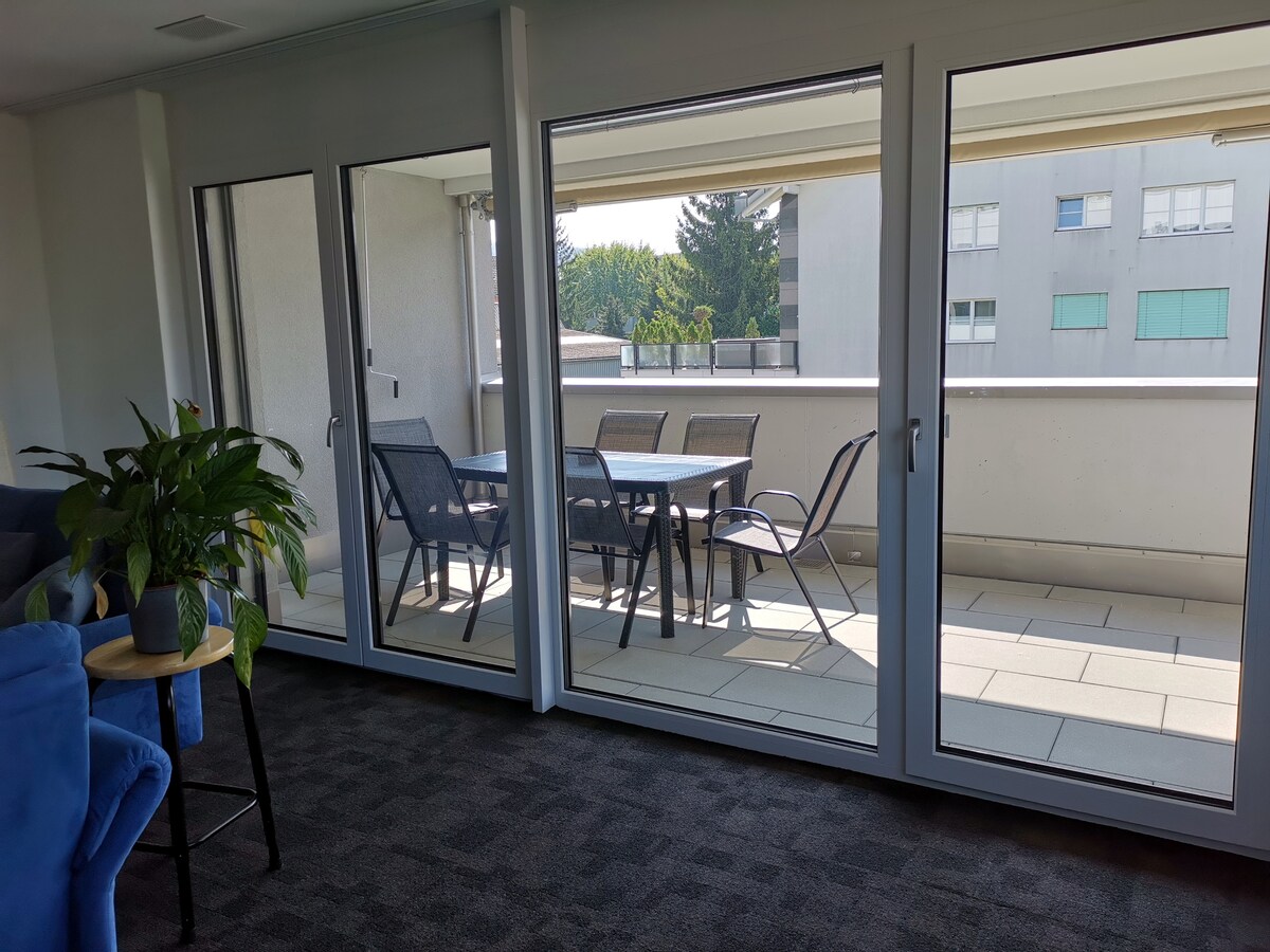 A bright living area features large glass doors leading to a spacious terrace. The terrace is furnished with a table and several chairs, surrounded by greenery. Natural light fills the space, highlighting the carpet and furnishings within the room.